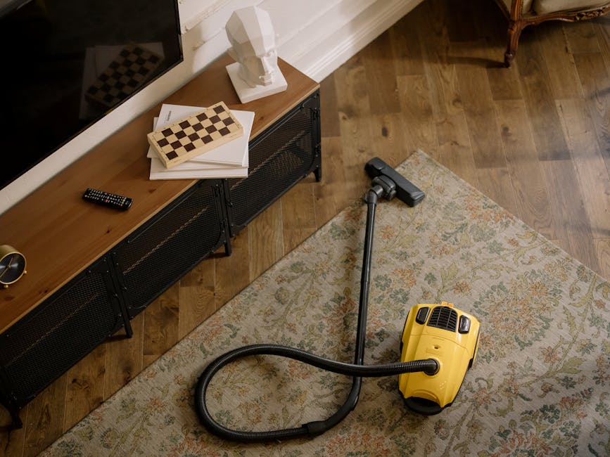 A yellow canister vacuum cleaner with a black hose and metal extension tube positioned on a patterned area rug in a living room. The room features wooden flooring and a modern black console table agai
