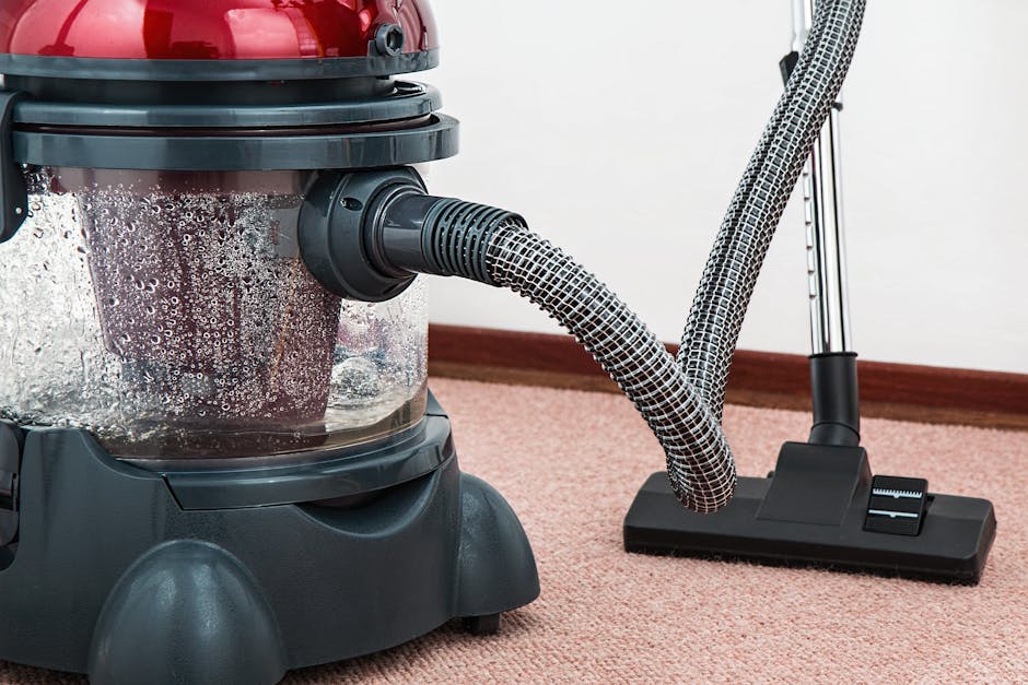 A yellow canister vacuum cleaner with a black hose and metal extension tube positioned on a patterned area rug in a living room. The room features wooden flooring and a modern black console table agai